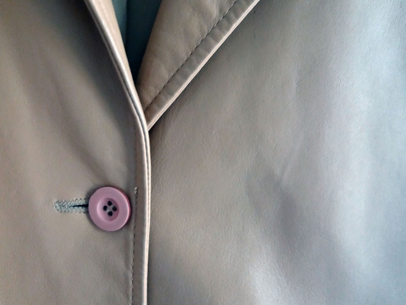 May include: A close-up of a beige leather jacket with a pink button.