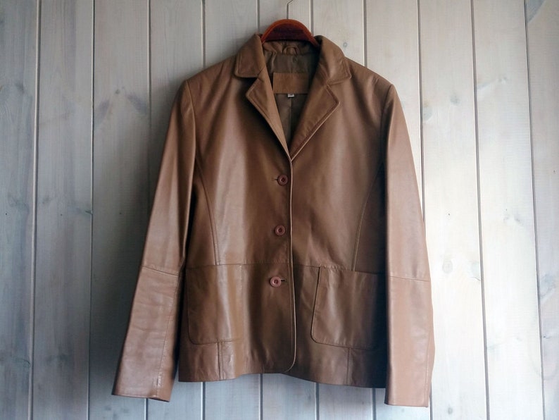 May include: A brown leather jacket with a button-up front and two pockets. The jacket is hanging on a wooden hanger.