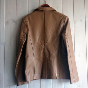 May include: A brown leather jacket with a zipper closure and a collar. The jacket is hanging on a wooden hanger against a light wood background.