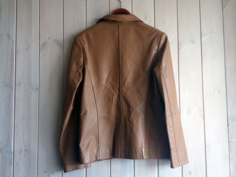 May include: A brown leather jacket with a zipper closure and a collar. The jacket is hanging on a wooden hanger against a light wood background.