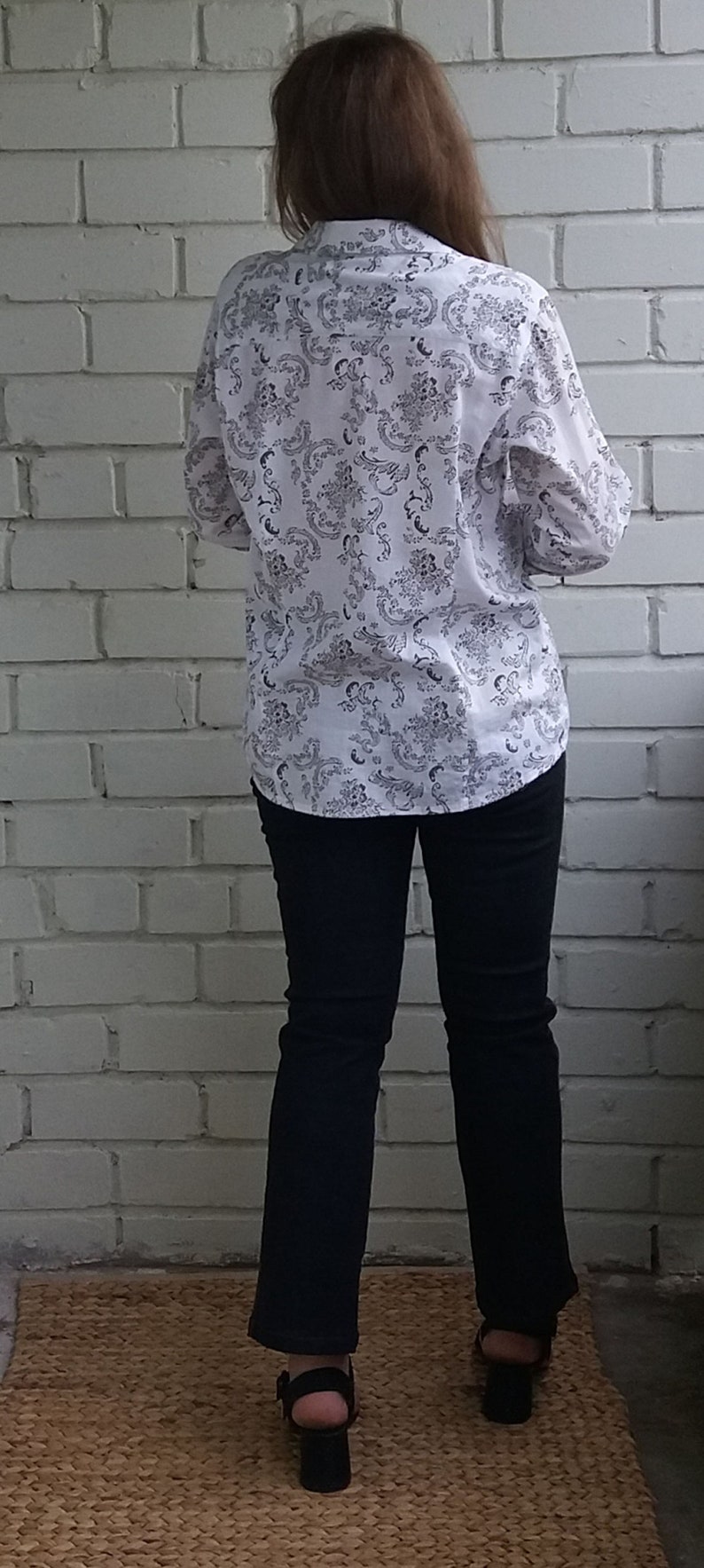 May include: A white button-down shirt with a black floral pattern. The shirt is worn with dark blue jeans and black heeled sandals.