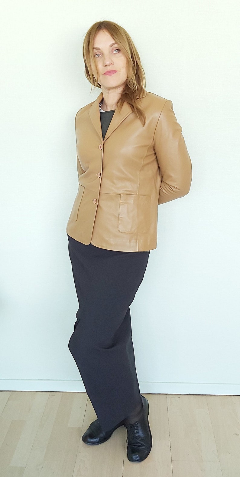 May include: A woman wearing a tan leather blazer with a black skirt and black shoes.