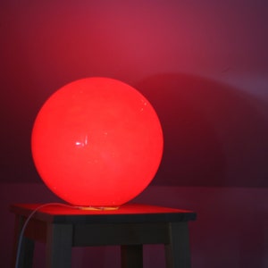 May include: A red spherical lamp sits on a wooden table. The lamp is lit and casts a warm glow on the surrounding area.