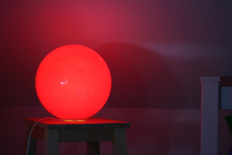 May include: A red spherical lamp sits on a wooden table. The lamp is lit and casts a warm glow on the surrounding area.