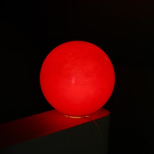 May include: A bright red sphere shaped lamp sits on a white surface. The lamp is lit and casts a warm glow.