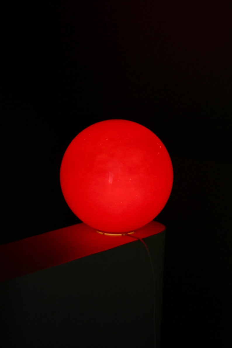 May include: A bright red sphere shaped lamp sits on a white surface. The lamp is lit and casts a warm glow.