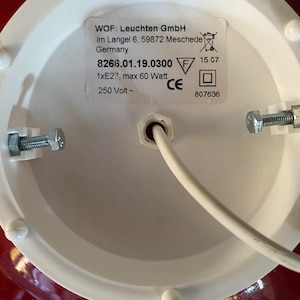 May include: Close-up of a white ceiling light fixture with a red base. The label on the fixture reads "WOF Leuchten GmbH Im Langel 6, 59872 Meschede Germany 8266.01.19.0300 1xE27, max 60 Watt 250 Volt ~ 15 07 CE 807636".