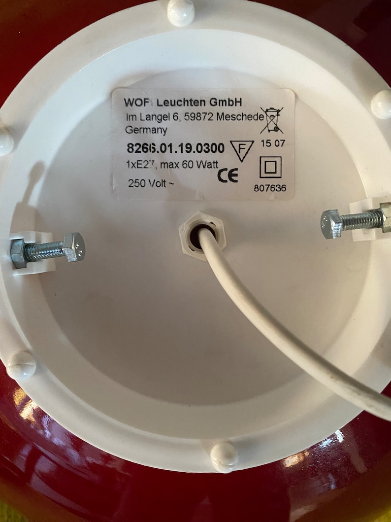 May include: Close-up of a white ceiling light fixture with a red base. The label on the fixture reads "WOF Leuchten GmbH Im Langel 6, 59872 Meschede Germany 8266.01.19.0300 1xE27, max 60 Watt 250 Volt ~ 15 07 CE 807636".