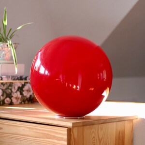 May include: A bright red, spherical lamp sits on a wooden dresser. The lamp is made of a smooth, glossy material and reflects light.