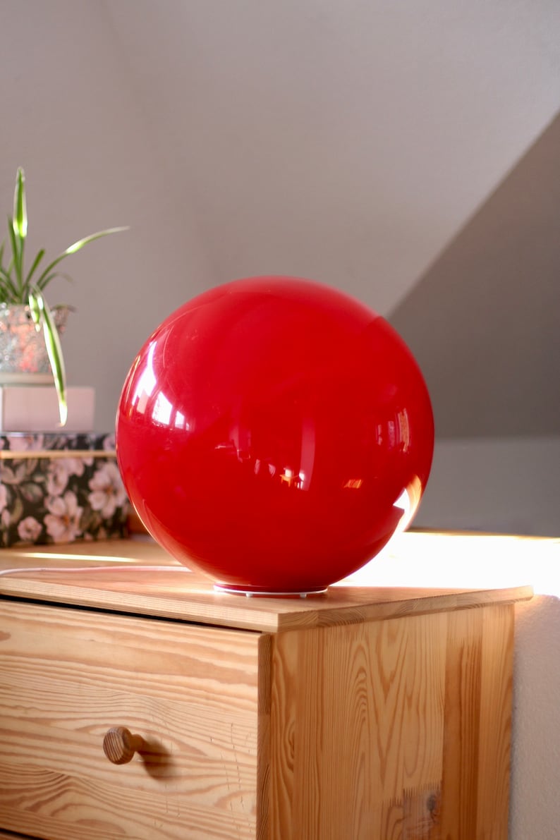 May include: A bright red, spherical lamp sits on a wooden dresser. The lamp is made of a smooth, glossy material and reflects light.