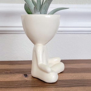 Puede incluir: Maceta de cerámica blanca con forma de persona sentada en posición de meditación, con una planta de aire verde creciendo dentro de la cabeza de la maceta.