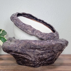 May include: A white ceramic planter with a textured, dark purple rim, nestled inside a larger, dark purple, textured ceramic planter that resembles a rock.