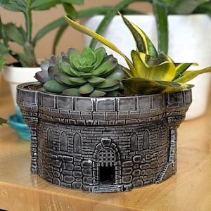 May include: A gray ceramic planter shaped like a castle tower, filled with green succulents and a yellow and green snake plant. The planter has a textured stone design with arched windows and a gated doorway.