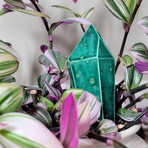 Handmade ceramic watering spike intricately shaped like a translucent green crystal with faceted edges, placed amidst the variegated green, white, and purple leaves of a Tradescantia plant in a white pot, offering a chic and eco-friendly plant care solution for indoor gardening enthusiasts.