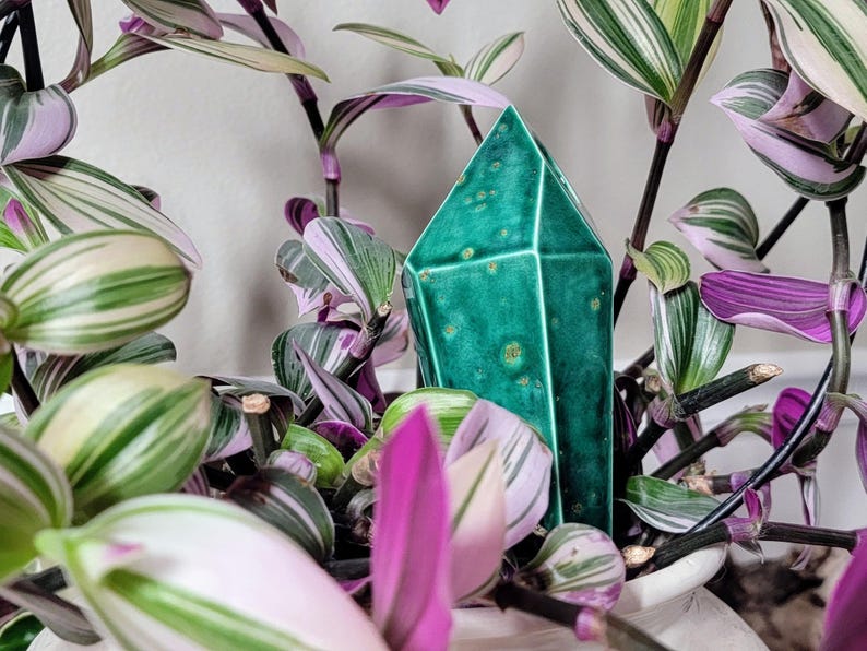 Handmade ceramic watering spike intricately shaped like a translucent green crystal with faceted edges, placed amidst the variegated green, white, and purple leaves of a Tradescantia plant in a white pot, offering a chic and eco-friendly plant care solution for indoor gardening enthusiasts.