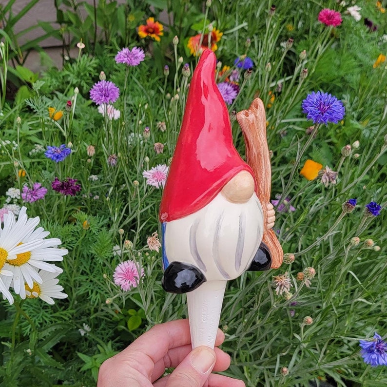Handmade ceramic watering spike featuring a whimsical garden gnome with a red hat, white beard, and black shoes, set against a vibrant background of blooming flowers including daisies, cornflowers, and multicolored blossoms, ideal for artistic and eco-friendly plant hydration.