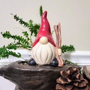 Handmade ceramic watering spike featuring a charming garden gnome with a red hat, white beard, and black shoes, holding a wooden stick, displayed against a dark stone-like surface with a pinecone accent and vibrant green fern plants in the background, blending functional hydration with whimsical decor for eco-conscious gardeners.