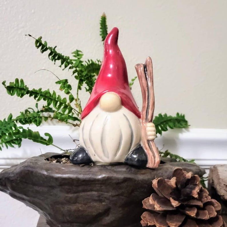 Handmade ceramic watering spike featuring a charming garden gnome with a red hat, white beard, and black shoes, holding a wooden stick, displayed against a dark stone-like surface with a pinecone accent and vibrant green fern plants in the background, blending functional hydration with whimsical decor for eco-conscious gardeners.