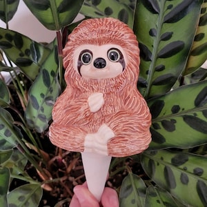 May include: A ceramic sloth planter with a brown and white fur design. The planter has a long, white spout for watering plants.