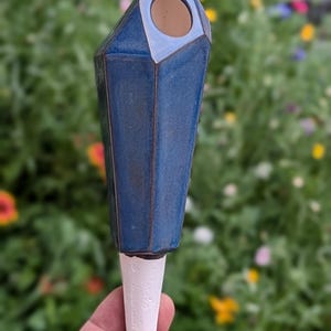Handmade ceramic watering spike with a faceted, geometric blue design and a white handle, featuring a circular opening for filling with water, photographed being held in a hand against a vibrant garden backdrop with lush foliage and colorful flowers, showcasing its eco-friendly functionality and stylish aesthetic for plant lovers.