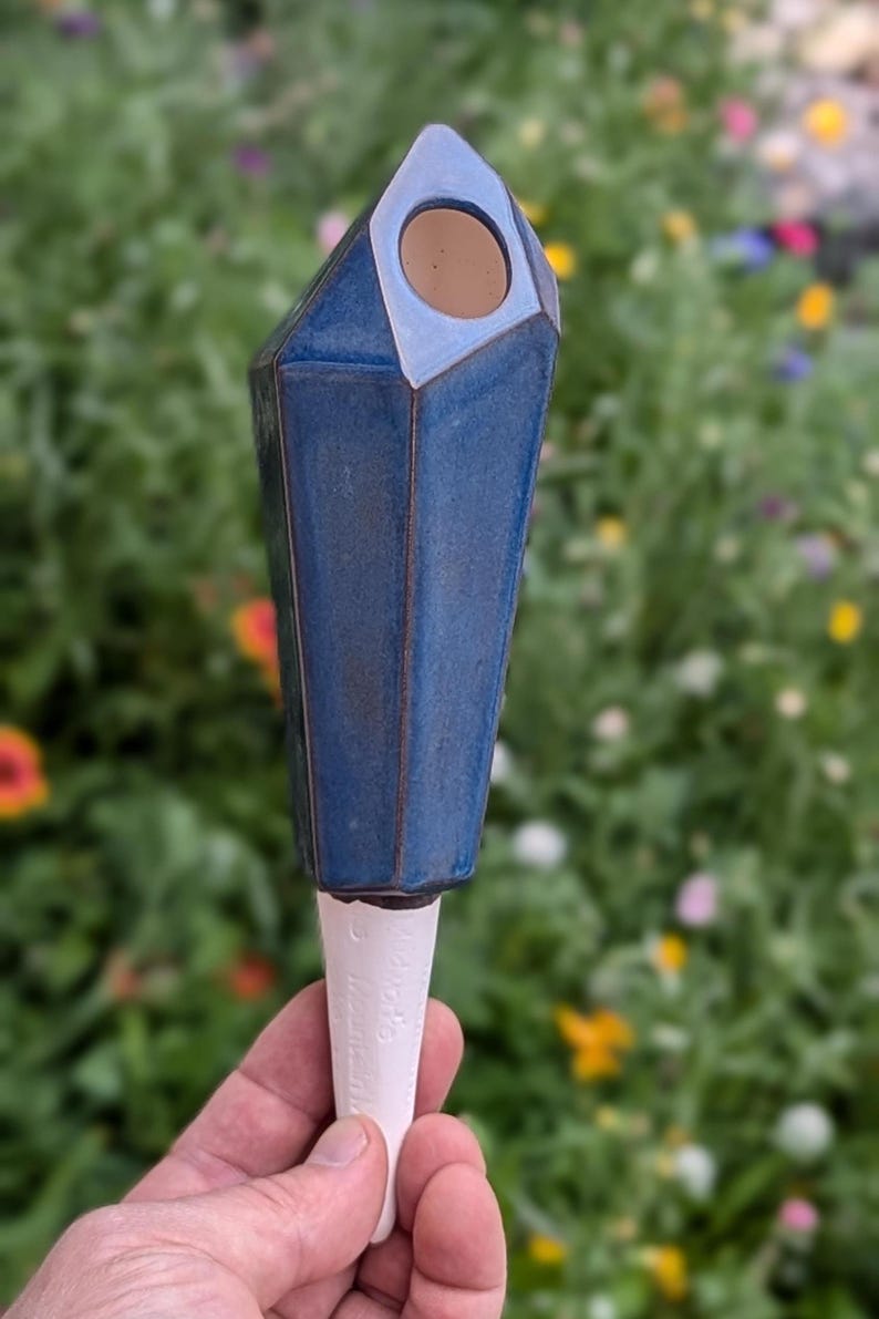 Handmade ceramic watering spike with a faceted, geometric blue design and a white handle, featuring a circular opening for filling with water, photographed being held in a hand against a vibrant garden backdrop with lush foliage and colorful flowers, showcasing its eco-friendly functionality and stylish aesthetic for plant lovers.