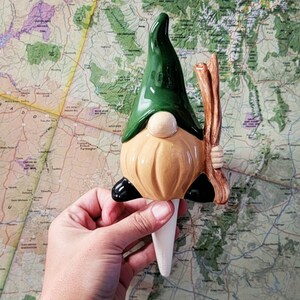 Handmade ceramic watering spike shaped like a garden gnome with a green hat, beige beard, and brown stick, displayed against a colorful topographical map background, blending charm and practicality for eco-conscious plant care enthusiasts.