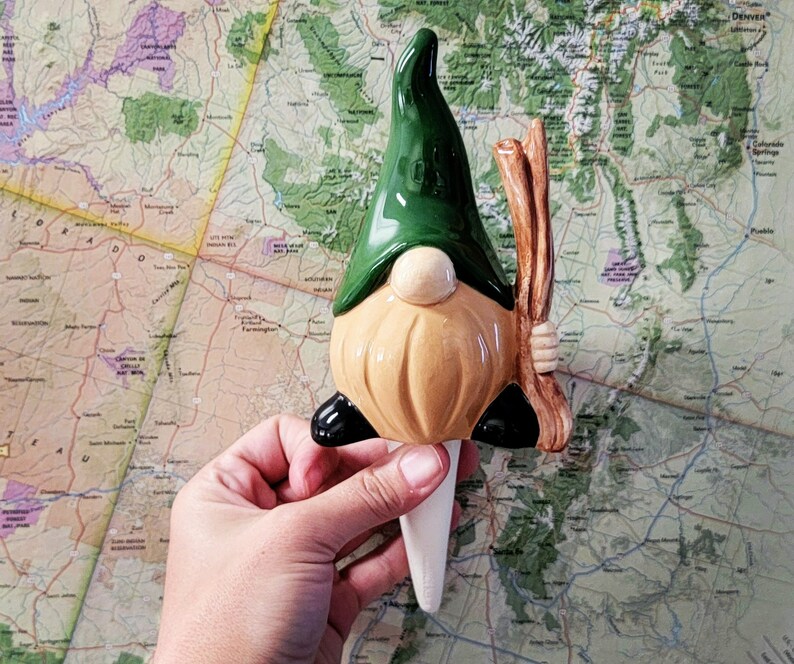 Handmade ceramic watering spike shaped like a garden gnome with a green hat, beige beard, and brown stick, displayed against a colorful topographical map background, blending charm and practicality for eco-conscious plant care enthusiasts.