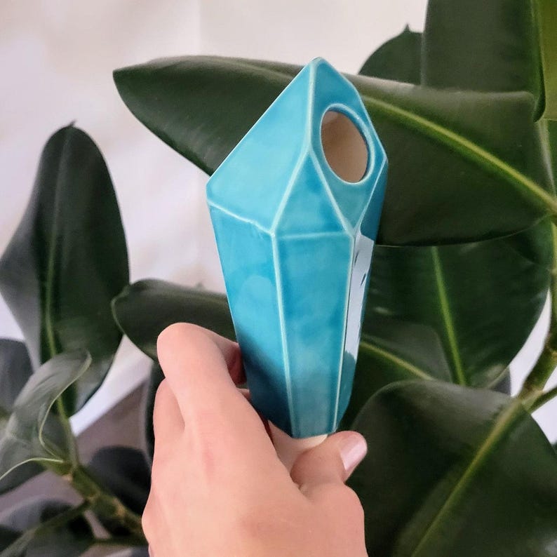 Handmade ceramic watering spike crafted in a geometric, faceted design resembling a vibrant blue crystal, featuring a small opening near the top for filling with water, held against a backdrop of lush green leaves from a rubber plant, ideal for eco-conscious plant enthusiasts.