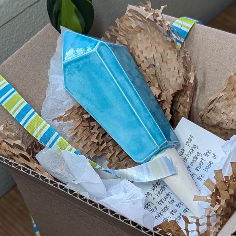 Handmade ceramic watering spike shaped like a vibrant blue crystal with angular facets, packaged in an eco-friendly cardboard box filled with brown crinkled paper and white tissue paper, accompanied by a striped ribbon and a handwritten note, emphasizing its functionality and sustainability for plant care enthusiasts.