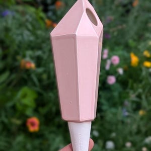Handmade ceramic watering spike in pink with a faceted, crystal-like design and a white spike embossed with "Mountain Mudworks" featuring a hole near the top for filling with water, held against a colorful garden backdrop with vibrant green plants and flowers, ideal for adding style and functionality to plant care routines.
