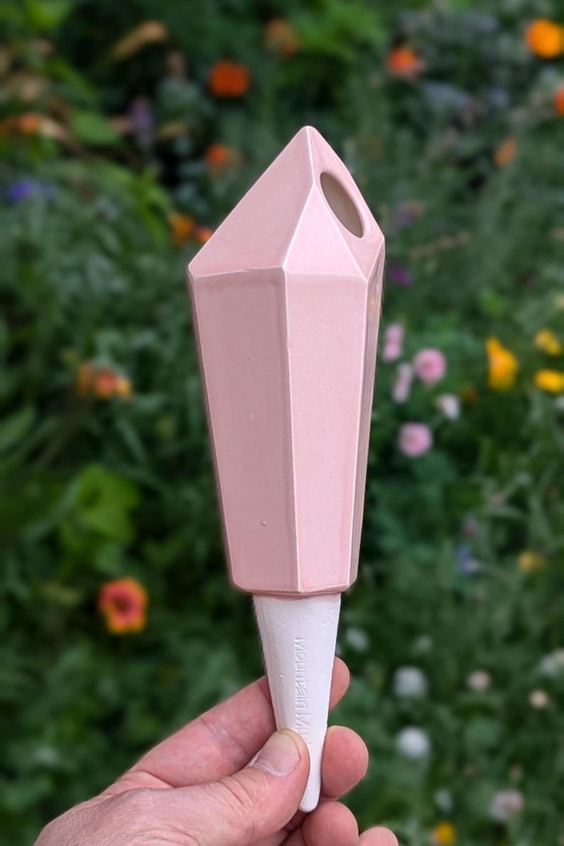 Handmade ceramic watering spike in pink with a faceted, crystal-like design and a white spike embossed with "Mountain Mudworks" featuring a hole near the top for filling with water, held against a colorful garden backdrop with vibrant green plants and flowers, ideal for adding style and functionality to plant care routines.