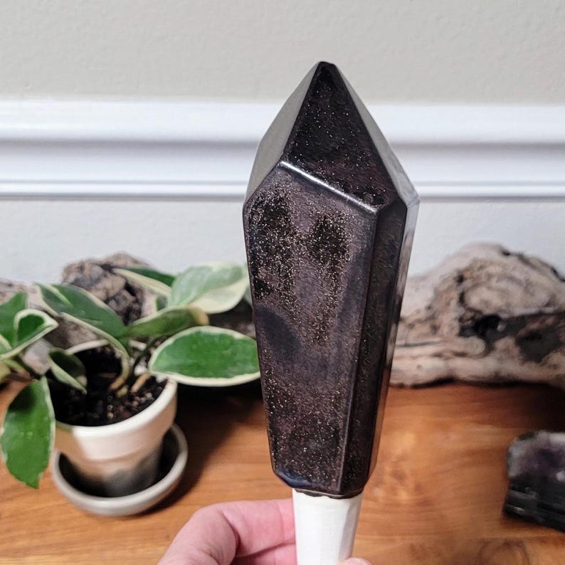 Handmade ceramic watering spike shaped like a dark crystal with sharp, faceted edges and a glossy, reflective surface, photographed with a potted plant featuring green and white variegated leaves and driftwood, set on a natural wooden surface indoors.