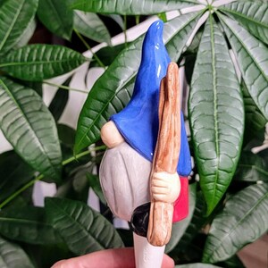 Handmade ceramic watering spike shaped like a whimsical garden gnome wearing a blue pointed hat, a red shirt, and white pants, positioned beside the green leaves of a houseplant, blending functional hydration and decorative charm for eco-conscious plant enthusiasts.