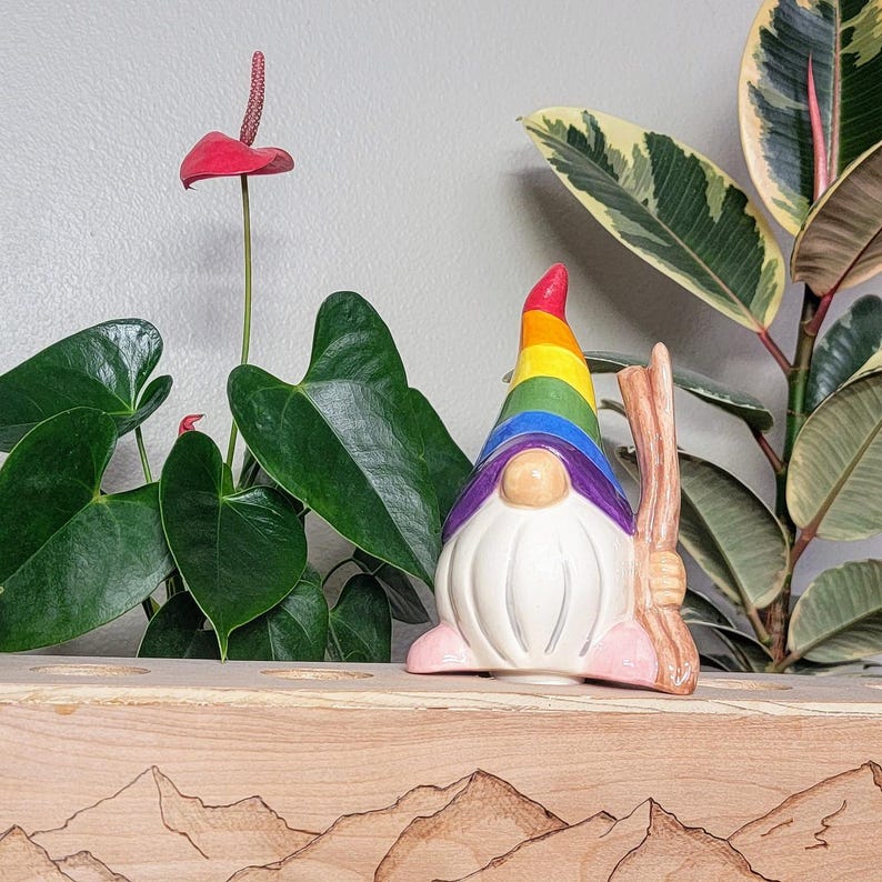 Handmade ceramic watering spike shaped like a gnome with a rainbow hat, placed among vibrant green houseplants including an Anthurium with a red flower and a variegated rubber plant. The gnome has a white beard, pink feet, and holds a wooden stick. The background features a wooden planter with engraved mountain designs.