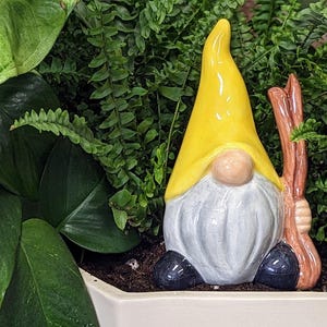 Handcrafted ceramic watering spike shaped like a garden gnome with a yellow hat, white beard, and a brown stick, nestled among large, vibrant green leaves in a white planter filled with soil, offering a whimsical and functional touch to plant care for eco-conscious gardeners.