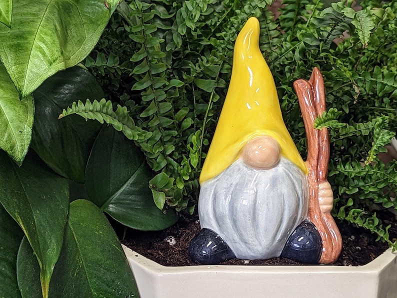 Handcrafted ceramic watering spike shaped like a garden gnome with a yellow hat, white beard, and a brown stick, nestled among large, vibrant green leaves in a white planter filled with soil, offering a whimsical and functional touch to plant care for eco-conscious gardeners.