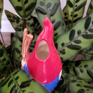 Handcrafted ceramic watering spike shaped like a gnome with a red hat, blue shirt, and white beard, holding a brown stick photographed from behind to show a round opening for filling and placed among the green spotted foliage of a houseplant, combining practicality and charm for eco-conscious plant enthusiasts.