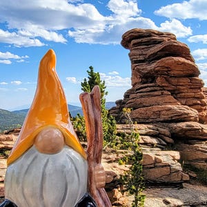 Handcrafted ceramic watering spike shaped like a garden gnome with an orange hat and white beard, holding a wooden stick, photographed against a scenic backdrop of layered rock formations, distant green mountains, and a blue sky with scattered clouds, combining whimsical design and natural aesthetics for eco-friendly plant care.