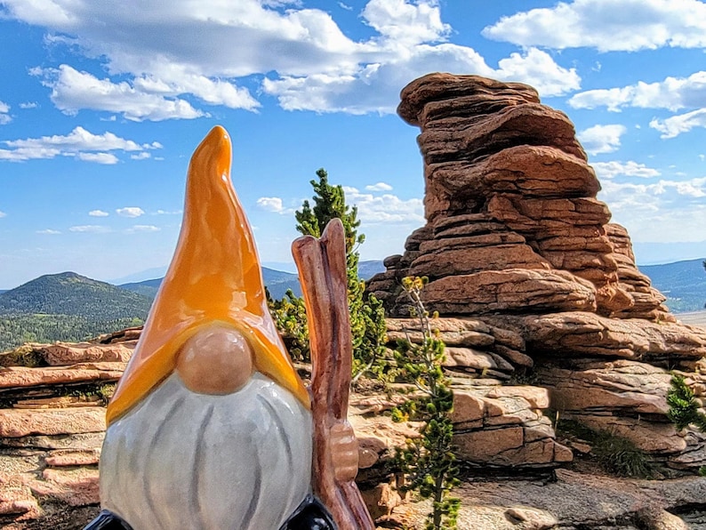 Handcrafted ceramic watering spike shaped like a garden gnome with an orange hat and white beard, holding a wooden stick, photographed against a scenic backdrop of layered rock formations, distant green mountains, and a blue sky with scattered clouds, combining whimsical design and natural aesthetics for eco-friendly plant care.