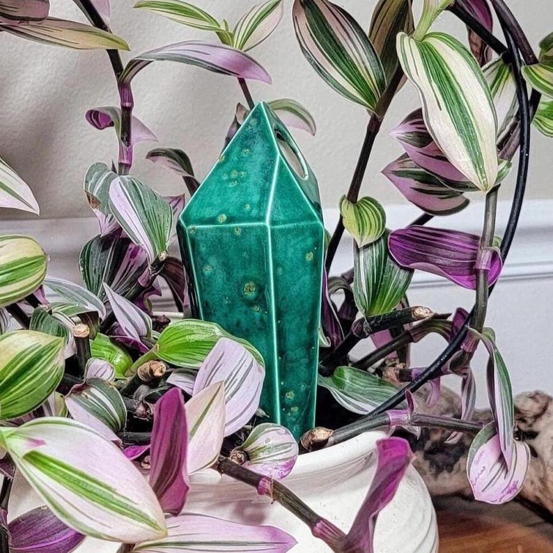 Handmade ceramic watering spike elegantly placed in a white pot, nestled among Tradescantia leaves displaying green, white, and purple variegation, complemented by a green crystal-like ornament in the center, photographed against a white wall and wooden surface.