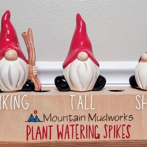 Handmade ceramic watering spikes shaped like whimsical garden gnome figurines, each featuring unique labels like “Hiking,” “Tall,” and “Short,” displayed on a rustic wooden block with the engraved artisan name “Mountain Mudworks,” accompanied by vibrant green foliage and a decorative glass jar.