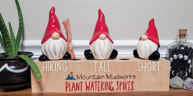 Handmade ceramic watering spikes shaped like whimsical garden gnome figurines, each featuring unique labels like “Hiking,” “Tall,” and “Short,” displayed on a rustic wooden block with the engraved artisan name “Mountain Mudworks,” accompanied by vibrant green foliage and a decorative glass jar.