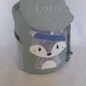 Könnte beinhalten: Ein grüner Rucksack mit Kordelzug und einem Fuchsgesicht auf der Vorderseite. Der Fuchs trägt ein blaues Stirnband und hat ein weißes Gesicht. Der Rucksack hat den Namen "Lani" auf der oberen Klappe gestickt.