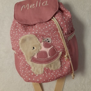 Könnte beinhalten: Rosa-farbener Rucksack mit weißen Punkten, einem Schildkröten-Applikationen und dem Namen "Melia" auf dem vorderen Klappdeckel.