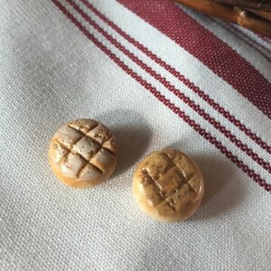 May include: Two small, round, brown and white bread rolls with a grid pattern on top. The rolls are on a white and red striped fabric.