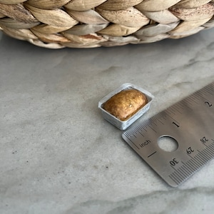 Puede incluir: Una hogaza de pan en miniatura en un recipiente blanco forrado de papel, que mide aproximadamente 2,5 cm de largo.