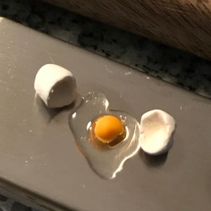 May include: A miniature white ceramic egg cracked open to reveal a yellow yolk and a clear egg white. The egg is sitting on a silver surface.