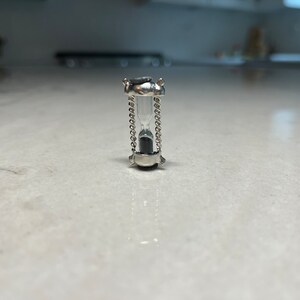 May include: A small silver hourglass with a clear glass bulb and black sand inside. The hourglass is on a white surface.