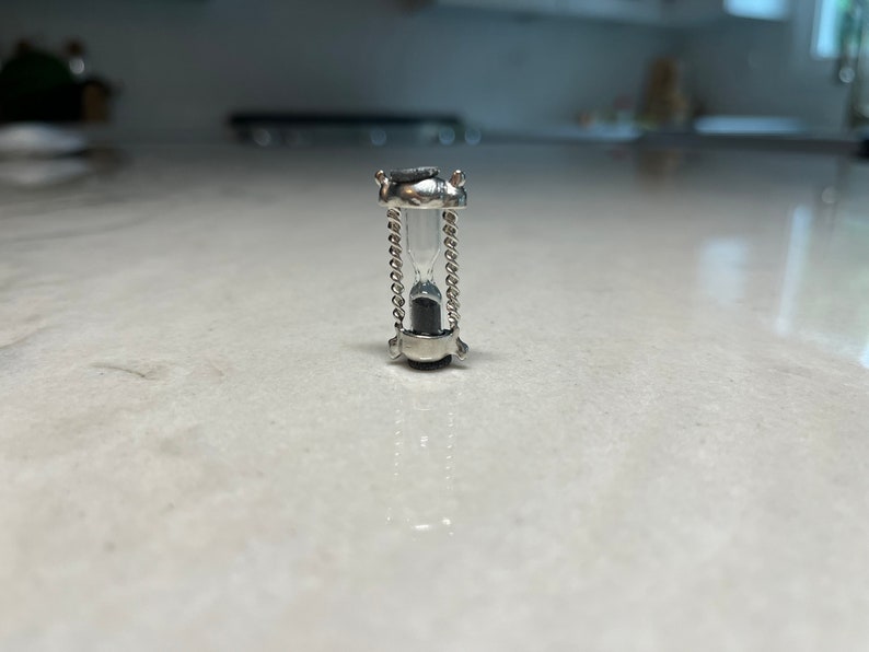May include: A small silver hourglass with a clear glass bulb and black sand inside. The hourglass is on a white surface.