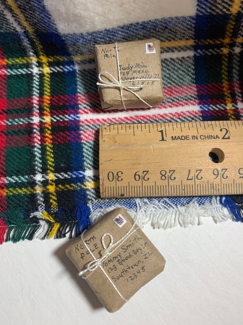 May include: Two small brown packages wrapped in twine with handwritten addresses to the North Pole. The packages are sitting on a plaid blanket with a ruler in the background.